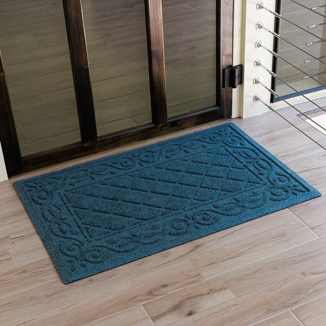 Angled top-down shot of a blue WaterHog Tristan mat resting just inside a dark wood-framed glass door. The natural light brings out the mat’s woven diamond texture and sculpted border, contrasting against the pale wood floor for a clean, refined entryway look.