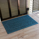 Angled top-down shot of a blue WaterHog Tristan mat resting just inside a dark wood-framed glass door. The natural light brings out the mat’s woven diamond texture and sculpted border, contrasting against the pale wood floor for a clean, refined entryway look.