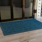 Blue WaterHog Tristan doormat placed just inside a dark wood-framed glass door, sitting on light wood floors with sunlight revealing its raised diamond-and-scroll texture and durable bi-level surface that catches dirt and moisture.
