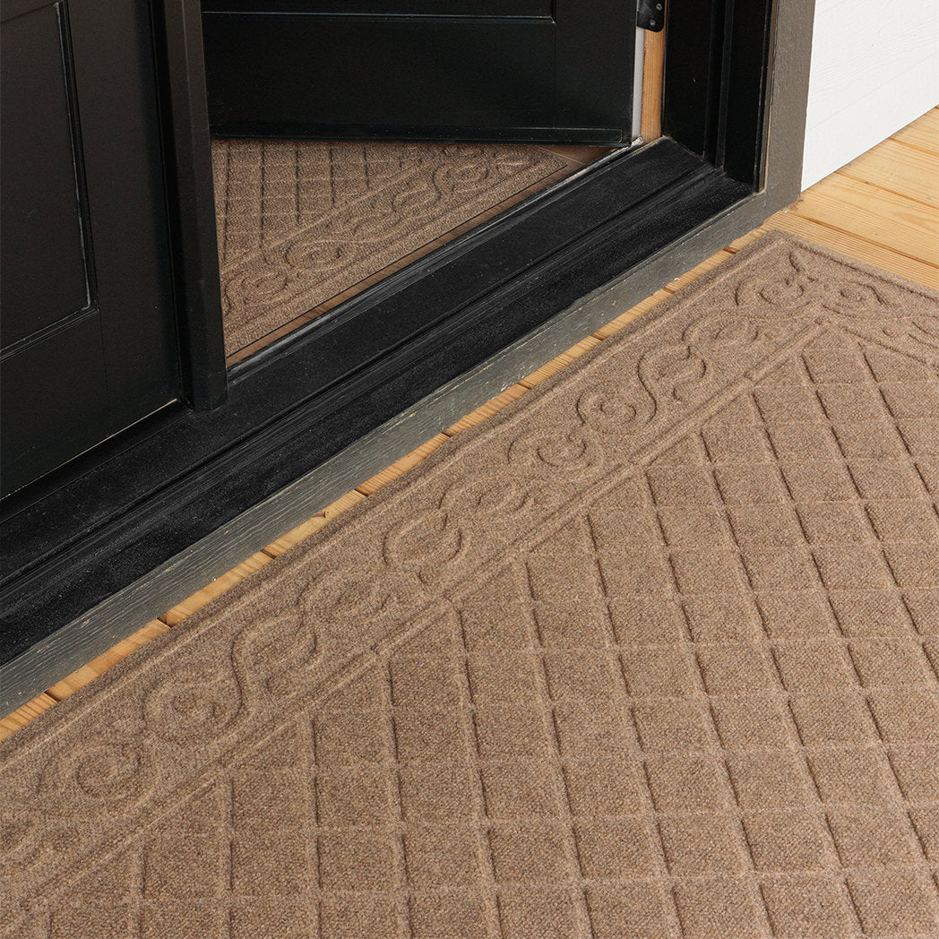 Angled close-up of two tan WaterHog Tristan mats at a black door—one runner inside and one doormat outside. The shot focuses on the raised scroll border and grid texture, showing how the matching mats frame the entryway against light wood floors.