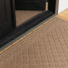 Angled close-up of two tan WaterHog Tristan mats at a black door—one runner inside and one doormat outside. The shot focuses on the raised scroll border and grid texture, showing how the matching mats frame the entryway against light wood floors.