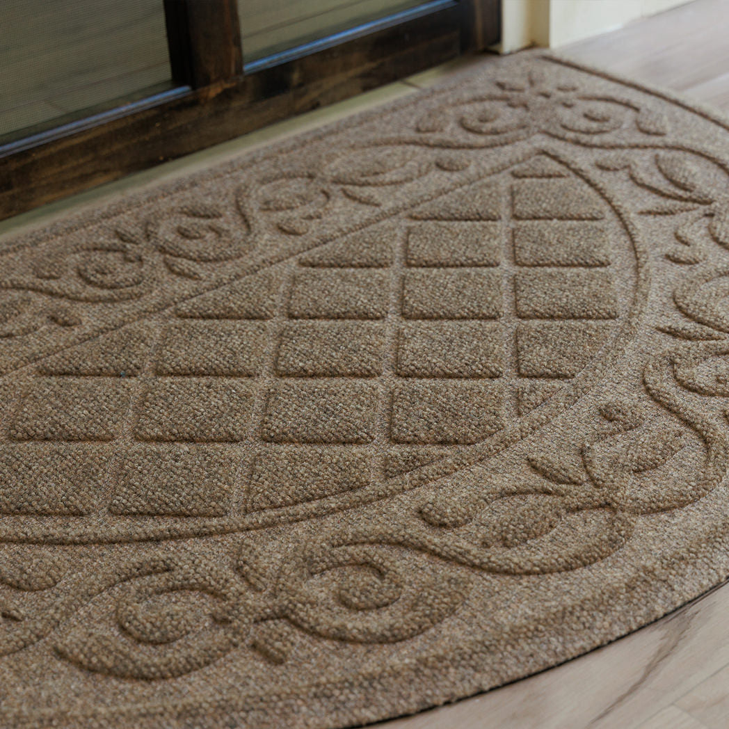 Tan WaterHog Tristan doormat with a rounded front edge placed at a wooden entry door, showing its raised diamond-and-scroll texture and bi-level surface that traps dirt and moisture from shoes.