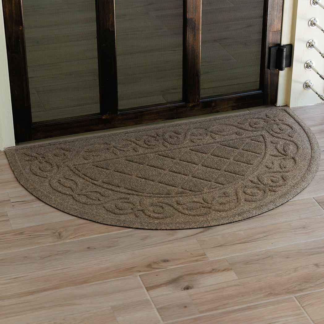Tan WaterHog Tristan doormat placed neatly against a wood doorway, its rounded front and textured scroll design adding a decorative touch while trapping mud and water.