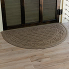 Tan WaterHog Tristan doormat placed neatly against a wood doorway, its rounded front and textured scroll design adding a decorative touch while trapping mud and water.