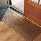 Tan WaterHog Tristan runner mat featuring an ornate raised pattern that traps dirt and moisture, shown here on light wood floors at an open entryway.