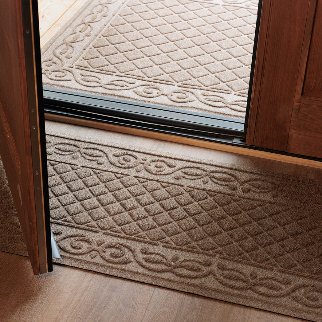 Tan WaterHog Tristan runner placed just inside a wood entryway and matching doormat outside the door, both featuring a raised diamond-and-scroll bi-level surface made of durable PET fibers that trap dirt and moisture on light wood flooring.