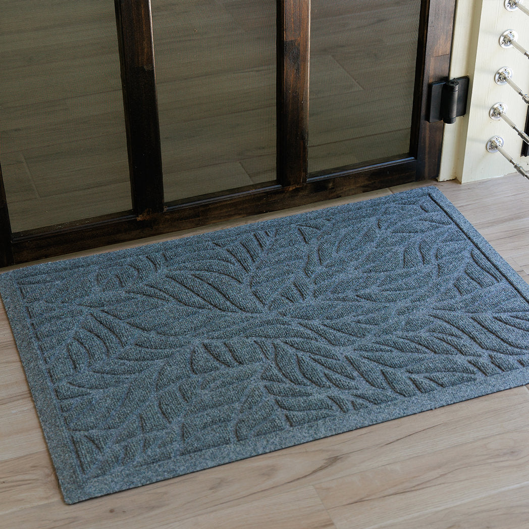 Blue-grey WaterHog Tropical Leaves doormat featuring a raised ridge design that traps dirt and moisture at an entryway on light wood floors.