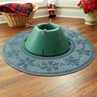 A green Christmas tree stand on a snowflake deco WaterHog Tree Skirt with holiday decor in the background.