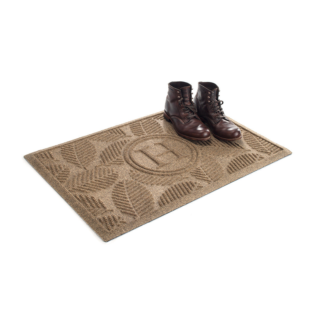 The OG WaterHog 2x3 Deanna monogram doormat in camel with boots placed on top on a white background.