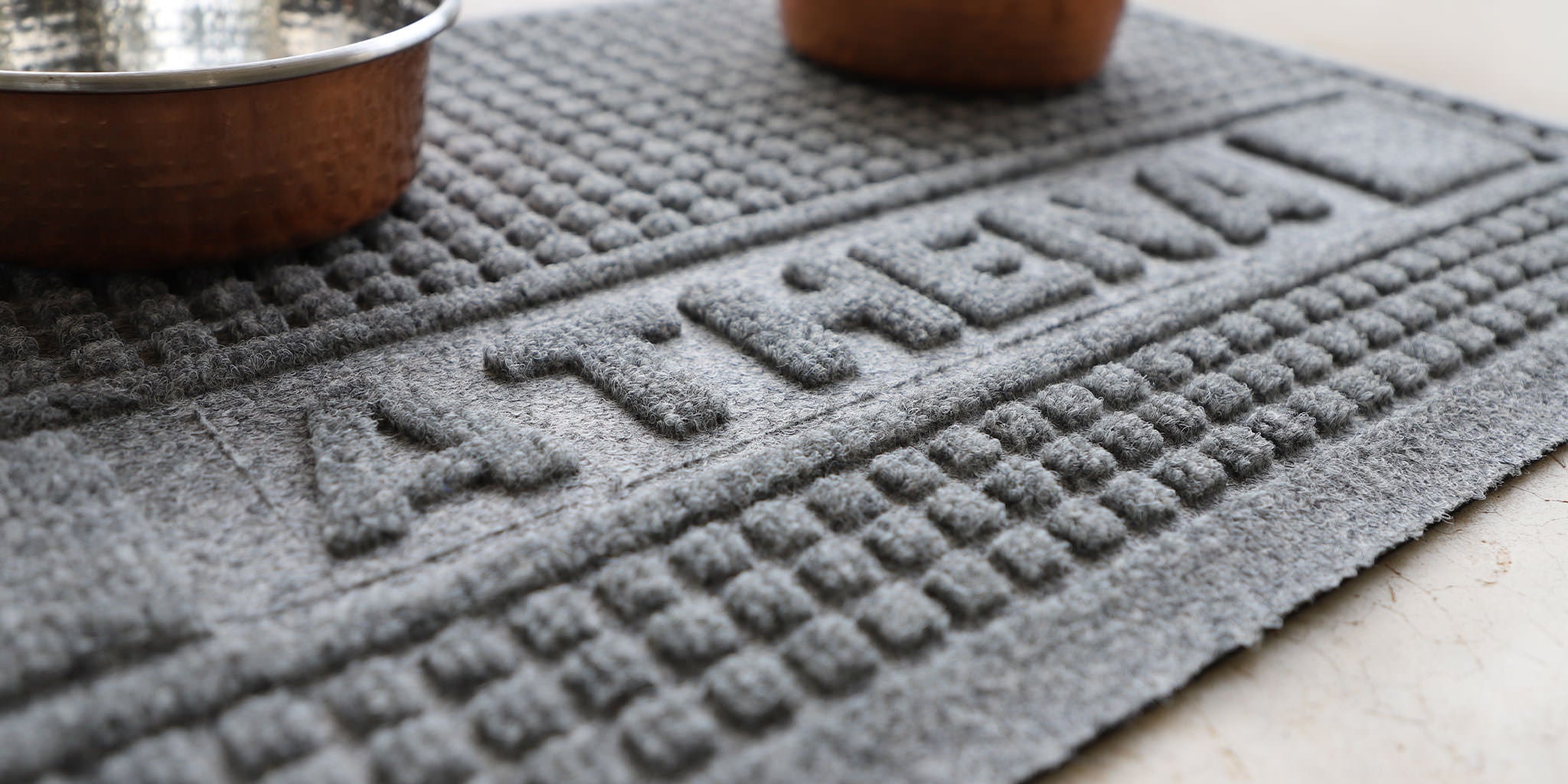 Zoomed in image of WaterHog Squares personalized pet bowl mat in a light grey color with the name Athena imprinted on it with pet bowls sitting on top. 