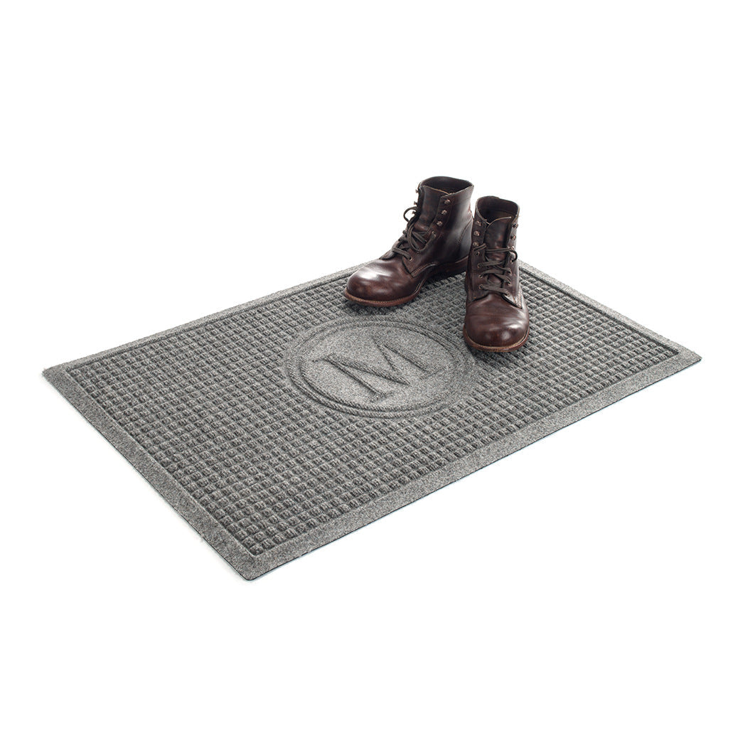 A pair of brown leather boots sitting on top of a WaterHog squares monogrammed mat in a light grey color with the letter 'M' in the center. 