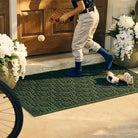 Young boy in baseball uniform with glove and ball, stepping out of his dirty cleats on a evergreen WaterHog canopy doormat. 