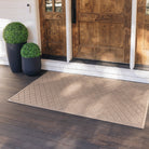 Extra large outdoor WaterHog doormat in a light brown color is placed at an all wood front door with large sidelights and flanked by green potted shrubs.