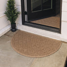 Unique half-round WaterHog outdoor doormat in classic light brown argyle pattern is placed on a stone front porch with a green potted shrub.