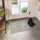 Floral themed WaterHog Boxwood indoor/outdoor doormat placed on stone color tile floors just inside a back door with pair of black boots on top; shown in medium grey color.