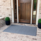 Entrance to a house with a wooden front door framed by narrow glass panels on either side. The flooring is made of light-colored brick with a large, rectangular, textured WaterHog cordova gray mat is placed in front of the door.