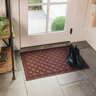 Indoor space near a glass door with white framing. The floor is tiled with gray stone tiles, creating a clean and modern look. A rectangular brown WaterHog doormat with a raised, decorative diamond pattern is placed in front of the door. A pair of black ankle boots is neatly positioned on the mat.