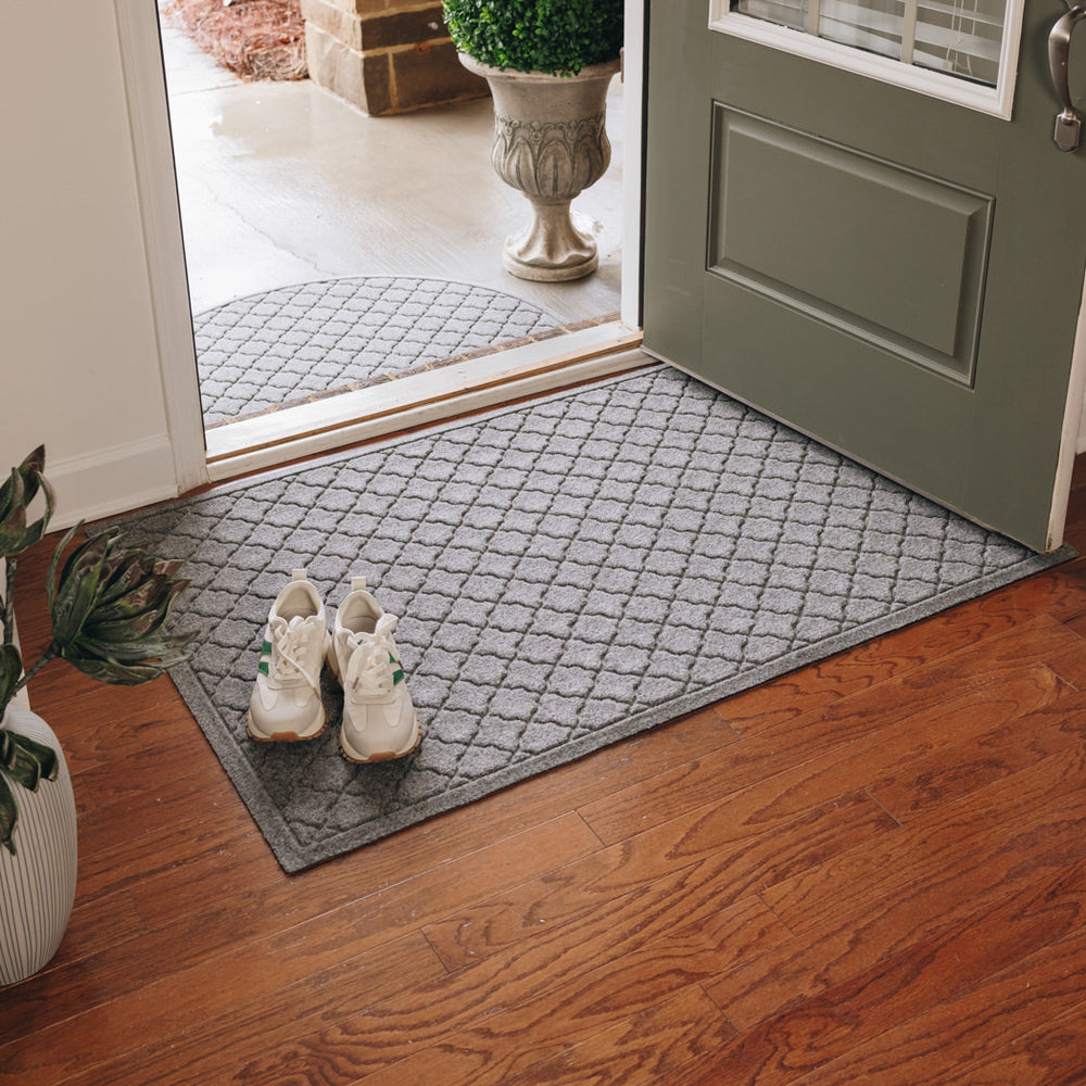 Open front door with a half-round WaterHog cordova in a medium grey on the outside of the door near wet pavement and a larger rectangular WaterHog mat in medium grey on the inside with a pair of sneakers sitting on top. 