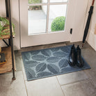 A medium-sized durable WaterHog doormat is placed inside in front of a single door that leads outside. The navy  blue doormat with a leafy design has a pair of black boots on top. 