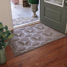 A large all-weather WaterHog doormat, in a tan color with a leafy design, is placed inside a single door leading to the outside. There is also a half-round outdoor doormat placed right outside the same door in a tan/golden color with a leafy design. 