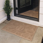 A medium-sized personalized WaterHog monogrammed all-weather doormat in a light tan camel color with a leafy design outside a single door on stone tile flooring. 