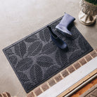 A medium-sized, durable WaterHog doormat in a dark gray/black color with a leafy design, placed outside on concrete flooring with a pair of navy blue boots with a few water spots on them. 