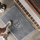 A medium grey monogrammed all-weather doormat, with a diamond design, is placed on concrete flooring; the feet of a person wearing white sneakers are shown standing on the doormat.  