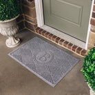 A medium-sized monogrammed WaterHog doormat in a medium grey color with a diamond pattern on concrete flooring placed in front of a single door. 