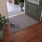 A light tan camel colored WaterHog doormat with a repeating dog bone pattern, sits on wood flooring inside a single door, with a dog leash resting on top; the door is open revealing a matching doormat outside, in the same color and pattern. 