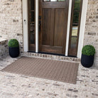 An extra-large outdoor mat in a light tan color with a repeating dog bone pattern placed on bricking flooring outside a single door with sidelights. 