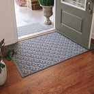 A large all-weather doormat in a light blue/grey color with an interlocking elliptical pattern sits on wood flooring inside a single door; the door is open revealing a medium-sized doormat outside, in the same color and pattern. 