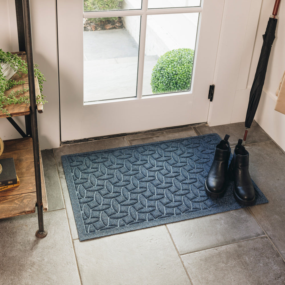 A medium-sized, navy blue durable WaterHog doormat with an interlocking elliptical design is placed on tile flooring inside a single door, with a pair of black boots sitting on top. 