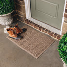 A tan WaterHog High Rise all-weather doormat with an elevated chevron-like design displayed in front of a door on concrete flooring.