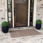 A brown WaterHog High Rise large doormat with an elevated chevron-like design displayed on brick flooring in front of a door.