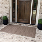 Extra-large WaterHog Lattice all-weather doormat in tan, featuring a crisscross geometric design, displayed on light brick flooring in front of a dark wooden front door.