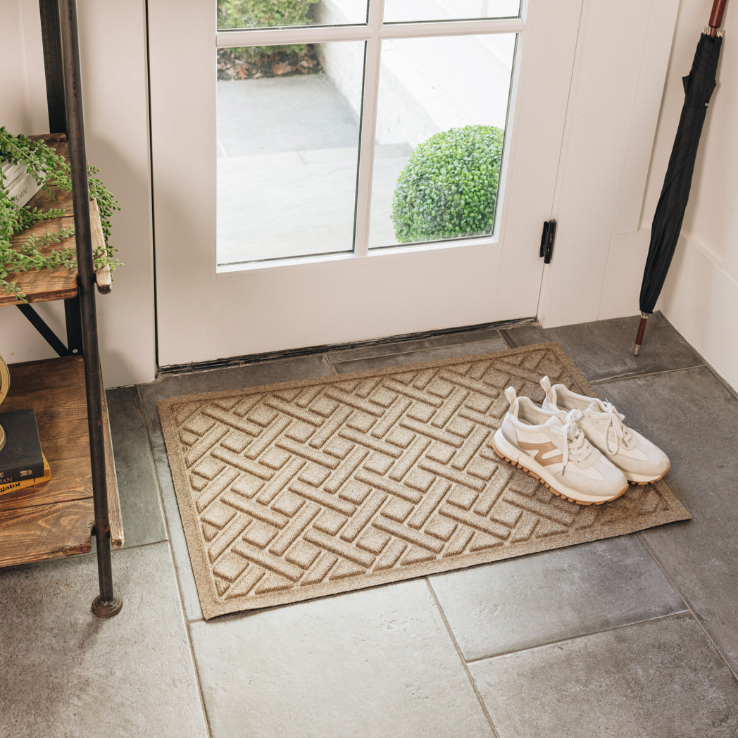 WaterHog Lattice indoor/outdoor mat in tan, featuring a crisscross design, displayed on grey tile flooring in front of a single door, with shoes placed on top.