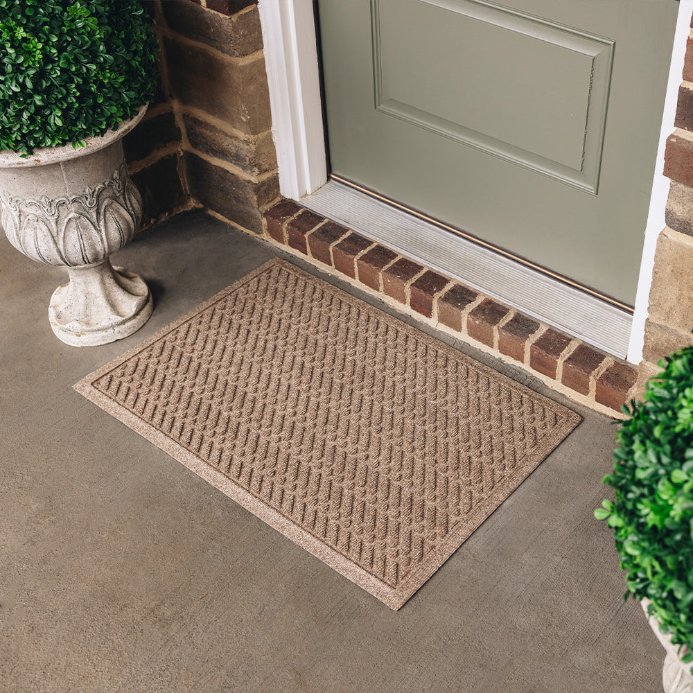 A WaterHog Sentry medium all-weather doormat in light tan, featuring a textured geometric pattern with diagonal ridges running in parallel lines, is displayed in front of a single door on concrete flooring.