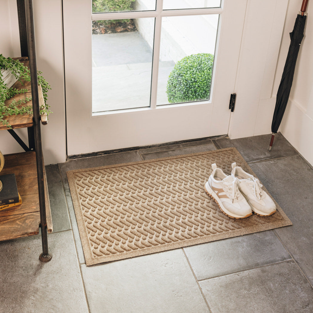 A WaterHog Sentry medium indoor/outdoor doormat in light tan, featuring a textured geometric pattern with diagonal ridges running in parallel lines, is displayed in front of a single door on tile flooring, with shoes placed on top of the mat.