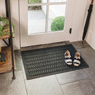 A WaterHog Sentry medium indoor/outdoor doormat in dark grey, featuring a textured geometric pattern with diagonal ridges running in parallel lines, is displayed in front of a single door on tile flooring, with shoes placed on top of the mat.