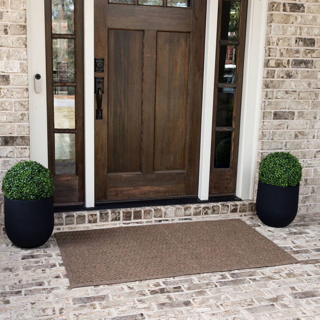 A WaterHog Sentry large indoor/outdoor doormat, featuring a textured geometric pattern with diagonal ridges running in parallel lines, is displayed in front of a single door on brick flooring.