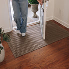 A WaterHog Sentry large indoor/outdoor doormat in light tan, featuring a textured geometric pattern with diagonal ridges running in parallel lines, is displayed on a wooden floor inside an entry with someone standing on the mat.