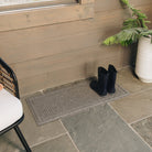 A grey WaterHog Boot Tray mat in the Squares design, on a stone porch with a pair of black boots drying off on the water-resistant, bi-level surface.