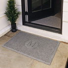 A WaterHog single door doormat in the traditional Squares design monogrammed with a serif M, at the side entrance on a stone ground.