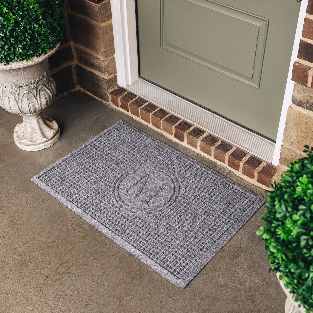 A grey WaterHog medium doormat, monogrammed in a center circle with the letter M, outside on a concrete entryway.