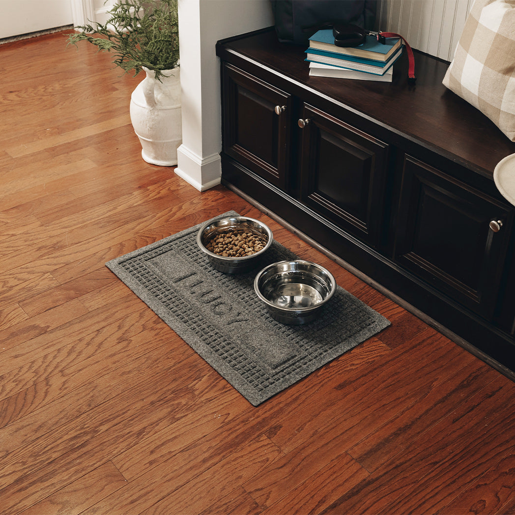 A medium WaterHog Squares pet mat personalized with the name LUCY, with a water and food dish on top while the mat protects the hardwood floors from spills and messes.