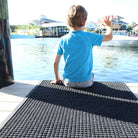 A young boy sitting and waving on a WaterHog Squares mat located at the edge of a dock.