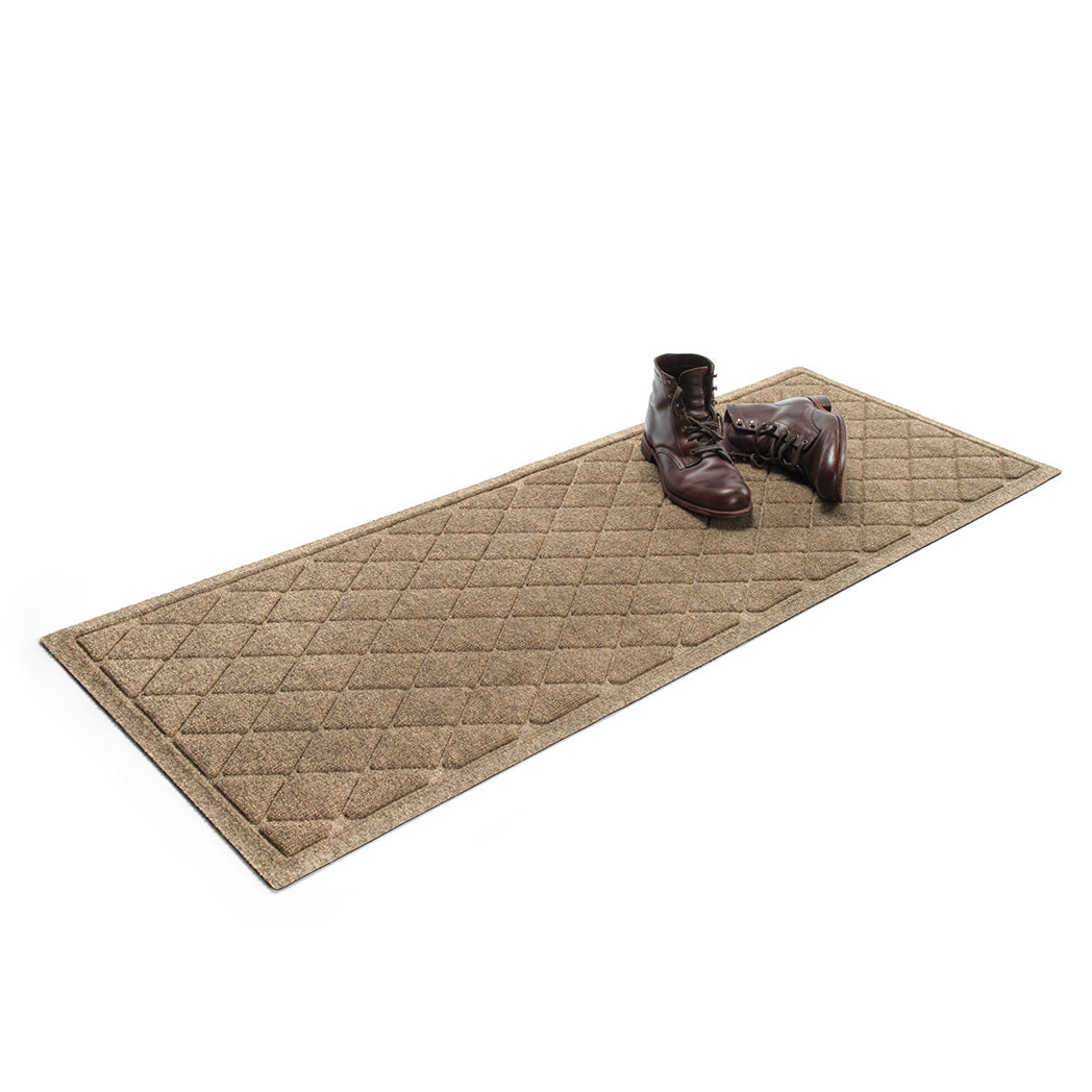 Isolated WaterHog 2x5 runner mat shown in a light tan with a pair of men's boots on top on a white background.