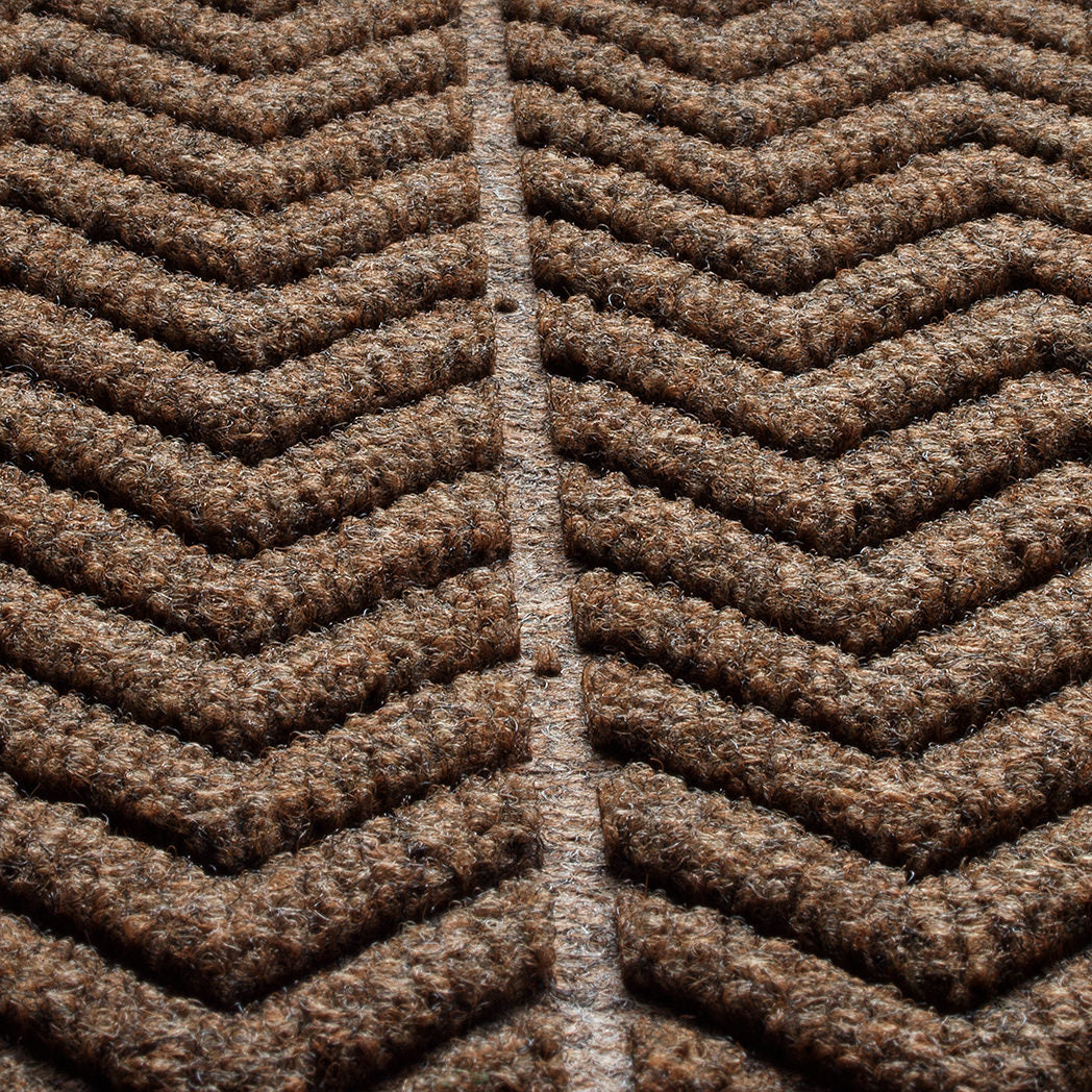 Close-up of a textured WaterHog doormat with a bold herringbone or chevron pattern made from a fibrous material consisting of cclors brown, black, and grey. The raised sections of the pattern create a three-dimensional effect designed for effective dirt and moisture trapping. 