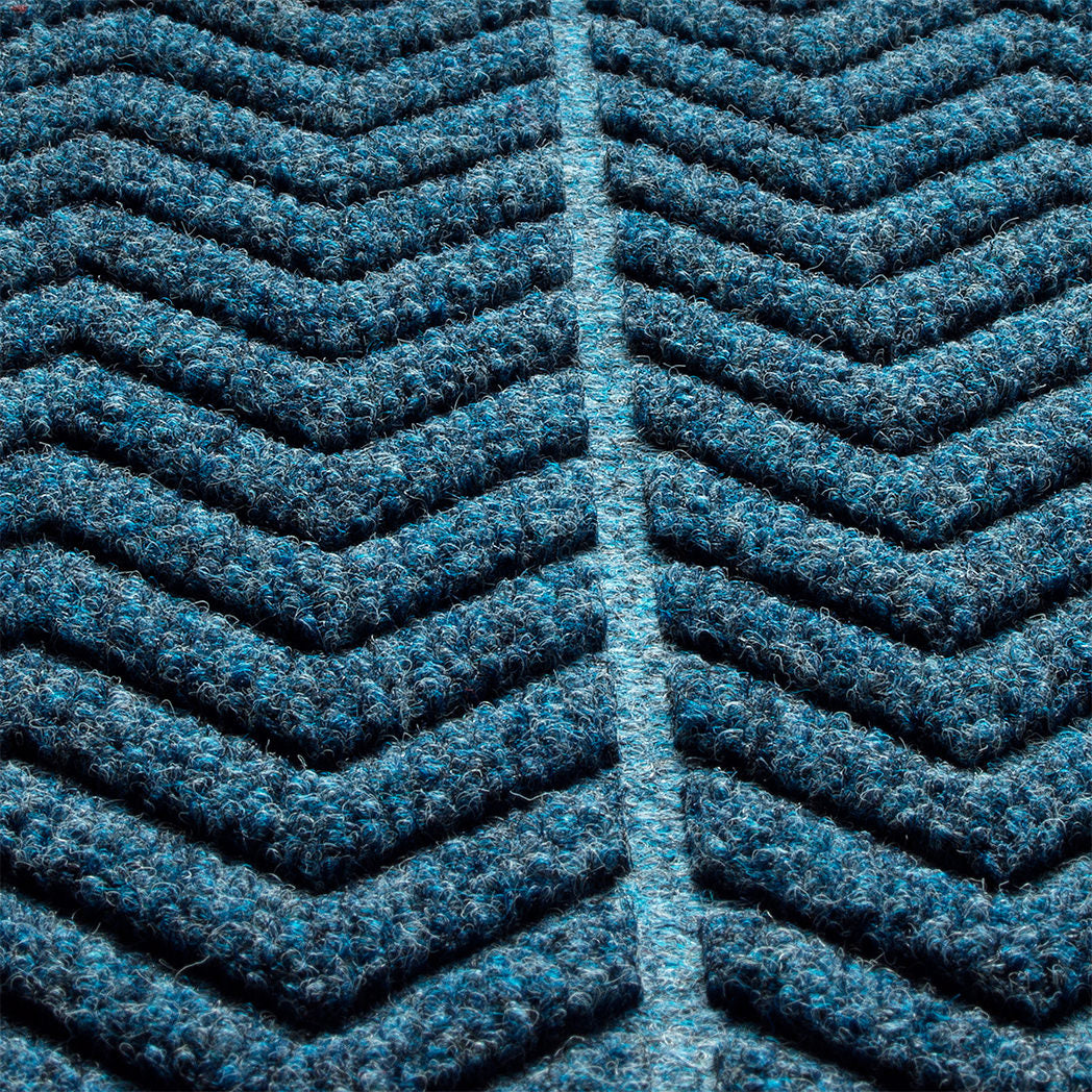 close-up of a WaterHog doormat with a chevron pattern. Made from a durable, recycled materials. The color scheme is predominantly deep blue with lighter blue highlights, giving it a rich, textured appearance. The raised sections of the pattern create a three-dimensional effect, suggesting that the mat is designed for effective dirt and moisture trapping, making it suitable for entryways or high-traffic areas.