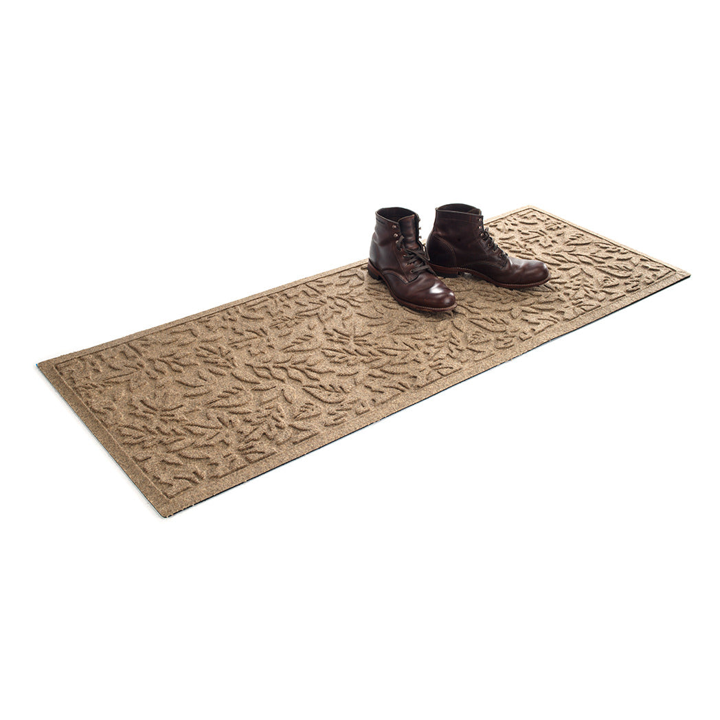 Isolated WaterHog 2x5 runner mat shown in a tan with a pair of men's boots on top on a white background.